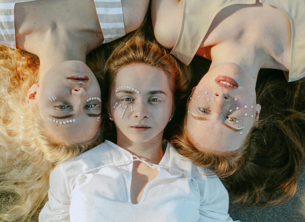 Tri devojke umetnice leže glavama oslonjene jedna na drugu, ukrašenih lica, mirnog pogleda. Autorka fotografije > pexels.com > Anfisa Eremina