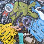 Bojana stoji ispred oslikanog zida i pruža ruke, a na zidu je mural oslikan apstraktnim motivima (linijama, bojama) i figurama plave ptice, rakuna i automata za loptice