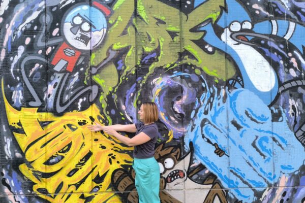 Bojana stoji ispred oslikanog zida i pruža ruke, a na zidu je mural oslikan apstraktnim motivima (linijama, bojama) i figurama plave ptice, rakuna i automata za loptice